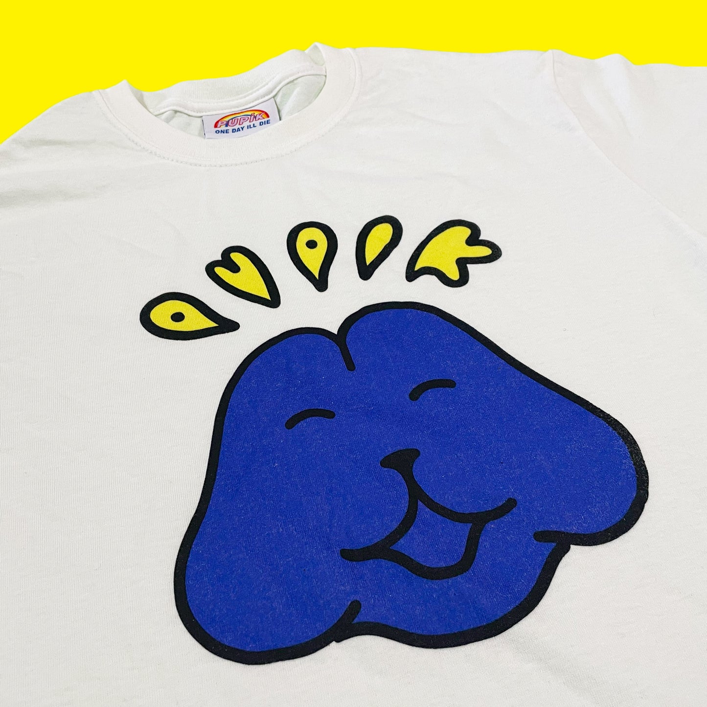 Pupik white T-shirt with a blue Pupik face graphic and yellow Pupik lettering printed on the front.