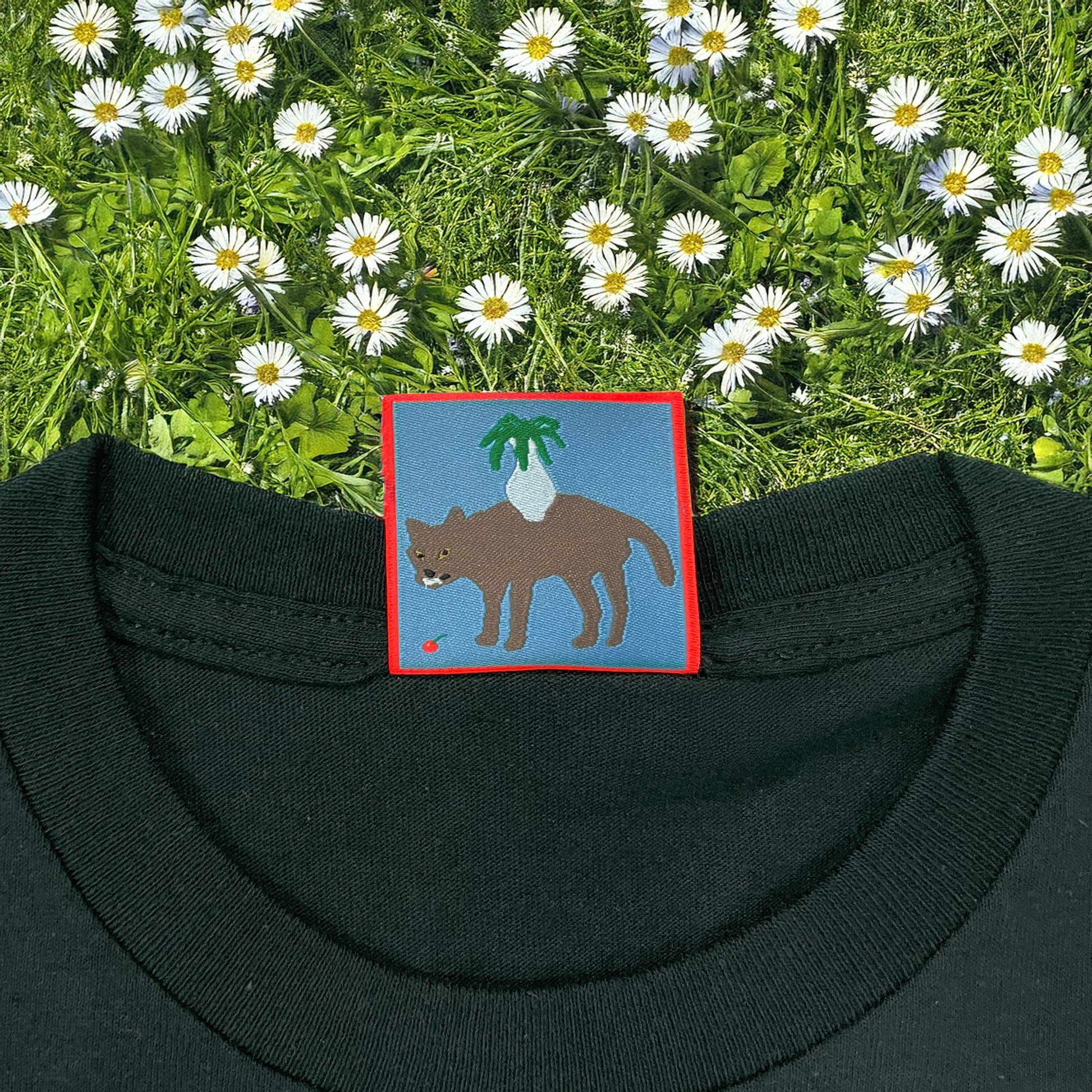 Close-up of Pupik Celebrating Perfection custom neck tag featuring an embroidered wolf with a radish-like plant on its back, framed in red on black fabric with daisies in the background