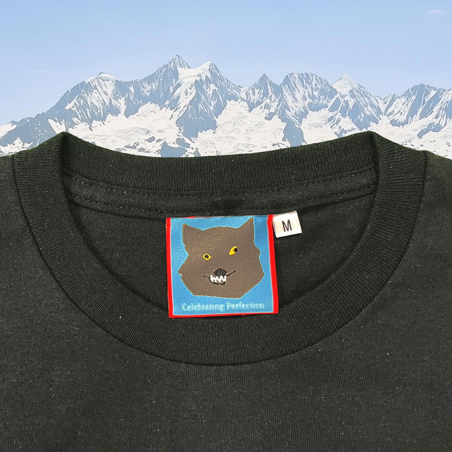 Close-up of Pupik Celebrating Perfection front neck tag featuring an embroidered wolf face with yellow eyes and sharp teeth on a blue background, framed in red with a mountain backdrop