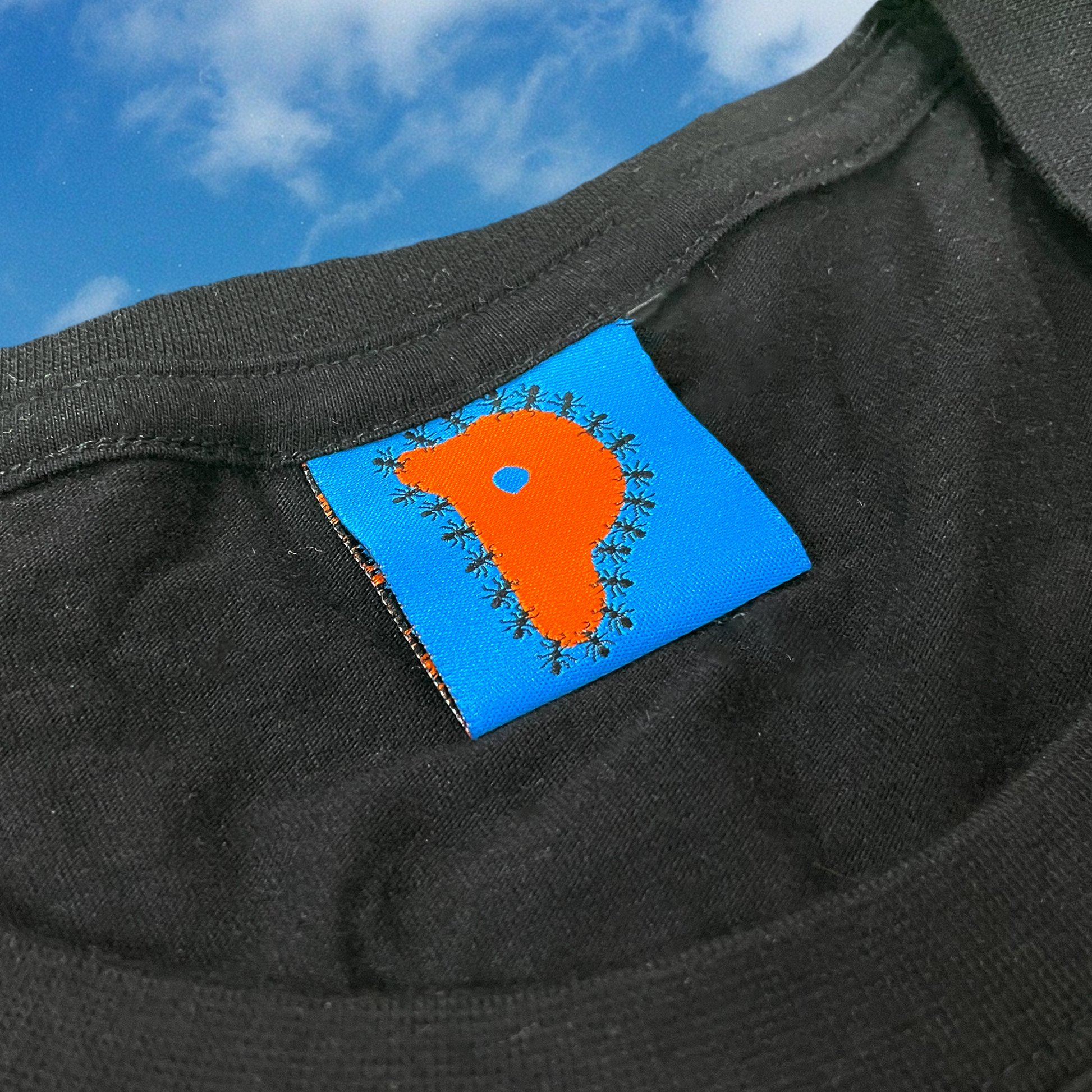 Close-up of Pupik woven tag on a black shirt, featuring an orange P surrounded by tiny black ants on a bright blue background, with part of the shirt collar visible against a cloudy sky backdrop