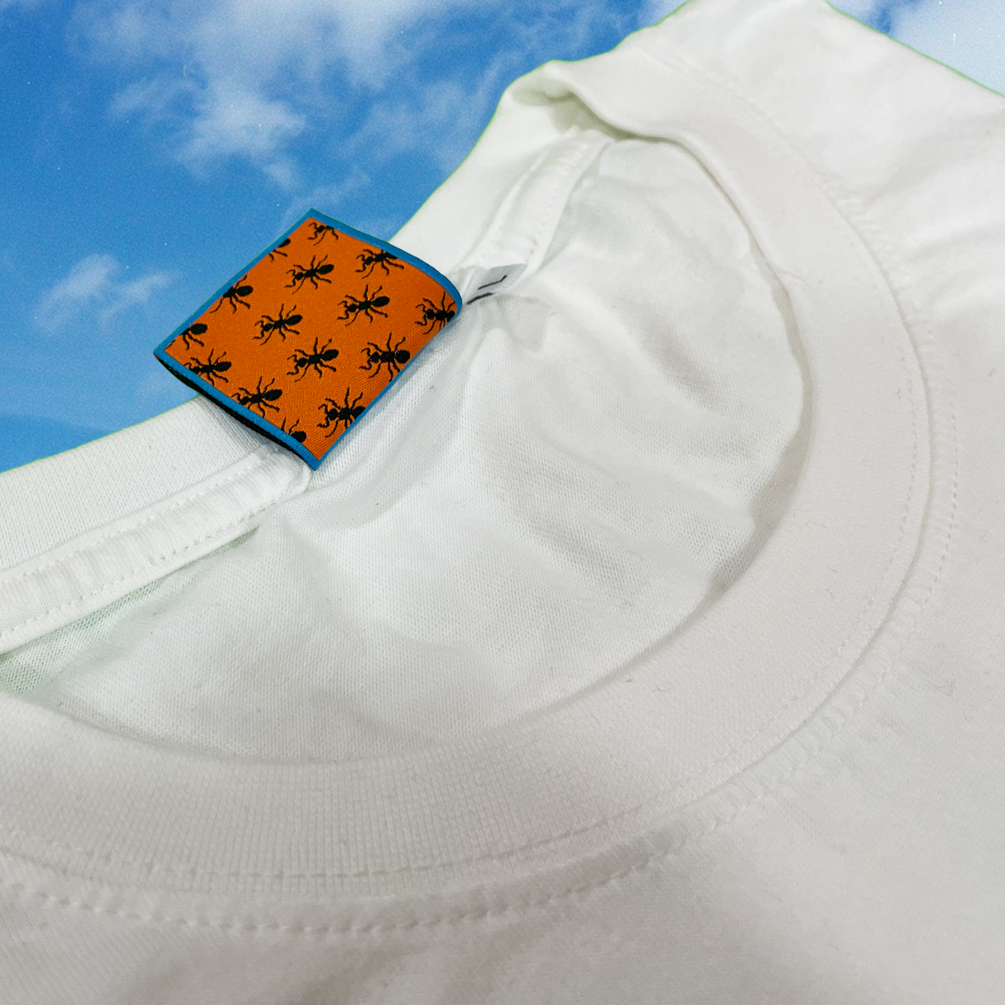 Close-up of Pupik woven tag on a black shirt, featuring an orange P surrounded by tiny black ants on a bright blue background, with part of the shirt collar visible against a cloudy sky backdrop