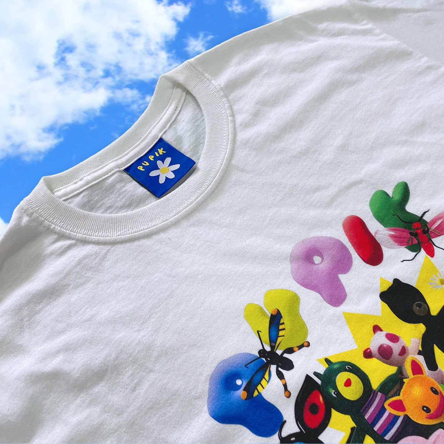 White t-shirt with colorful designs and a brand label against a blue sky.