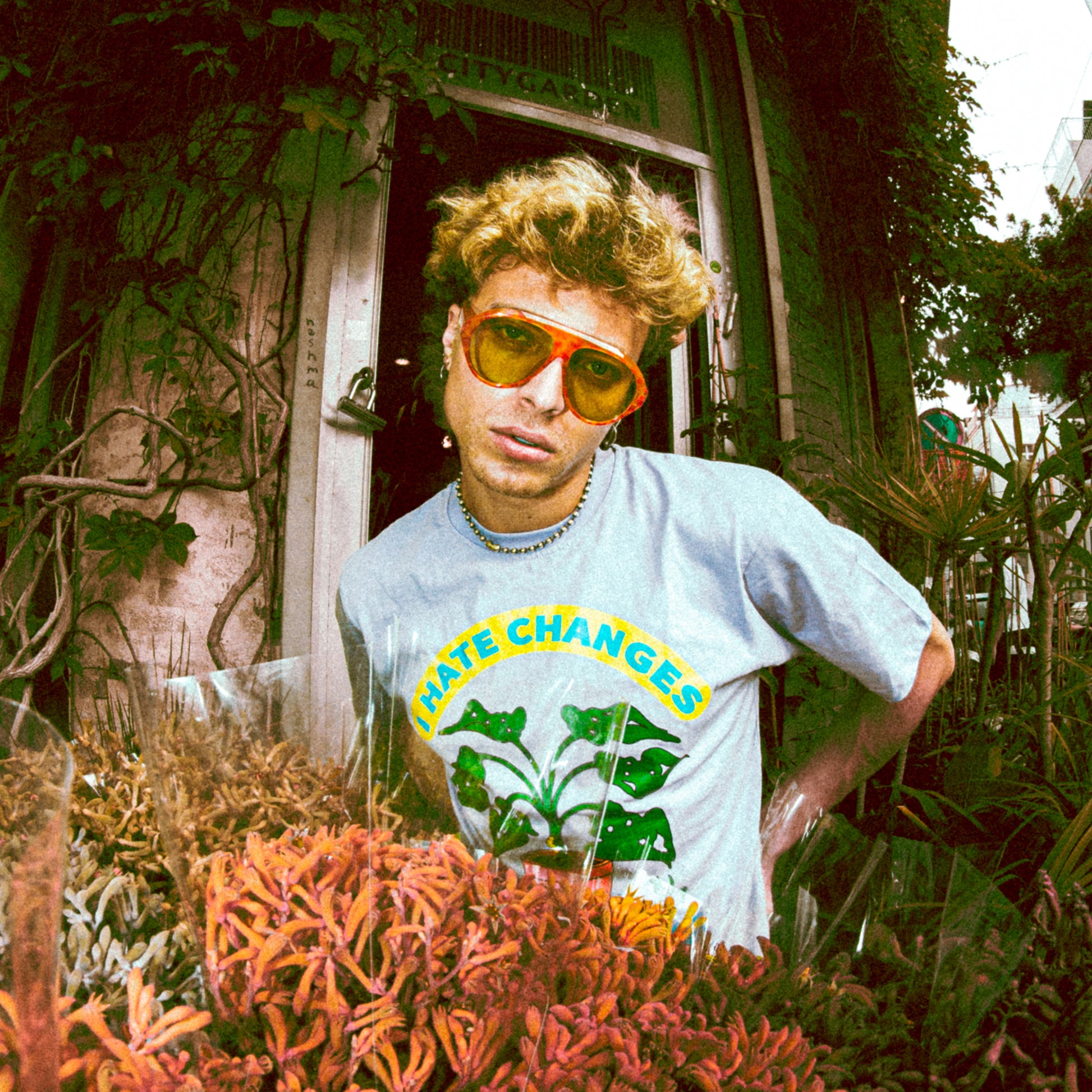 Person wearing a 'hATE CHANGES' t-shirt with sunglasses in front of a building with plants.